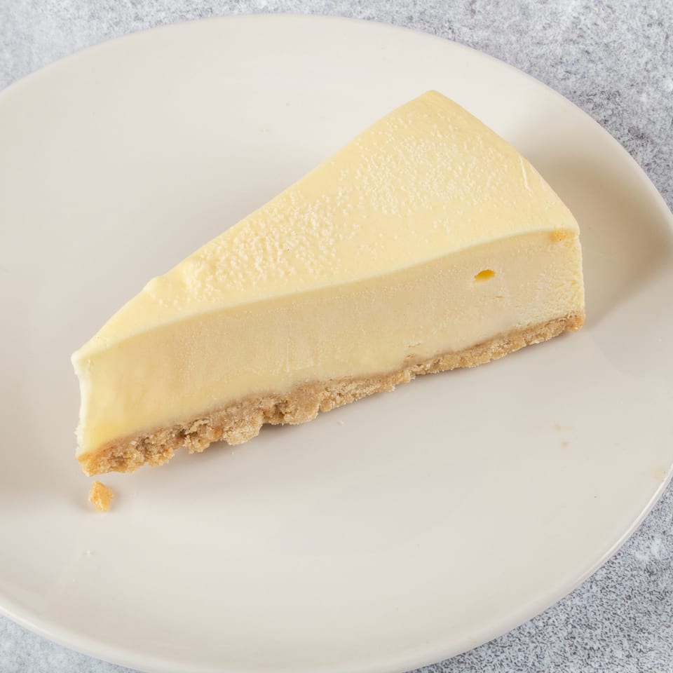 A slice of classic cheesecake with a graham cracker crust on a white plate.