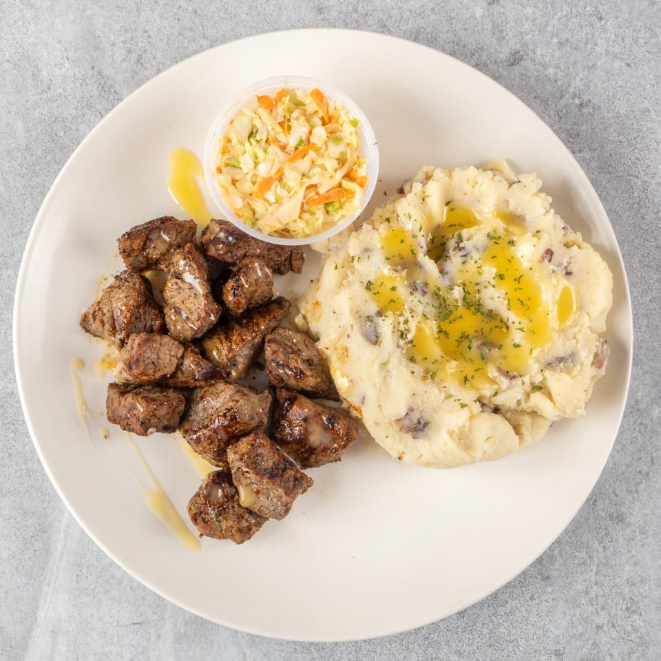Steak tips with mashed potatoes drizzled with butter and a side of coleslaw on a white plate.